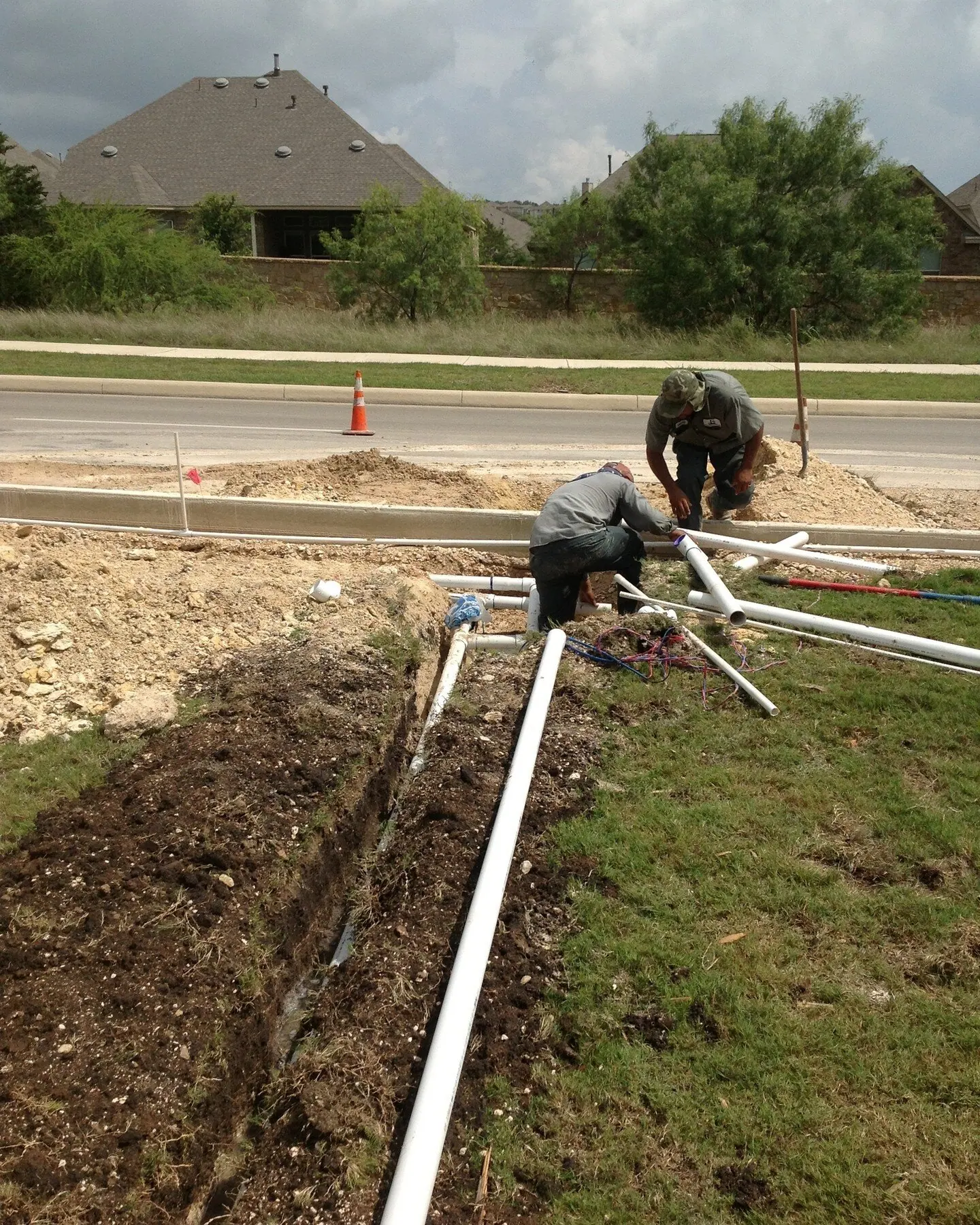 Two workers kneeling at a pipe trench beside a road